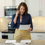 Professional woman in kitchen on the phone while looking at paperwork on the counter.